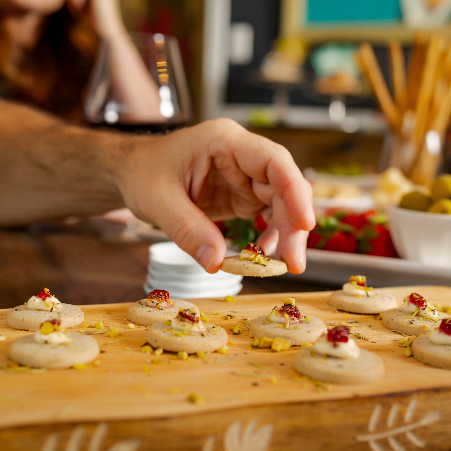
                  
                    Person preparing appetizers on a wooden board with various ingredients in the background
                  
                