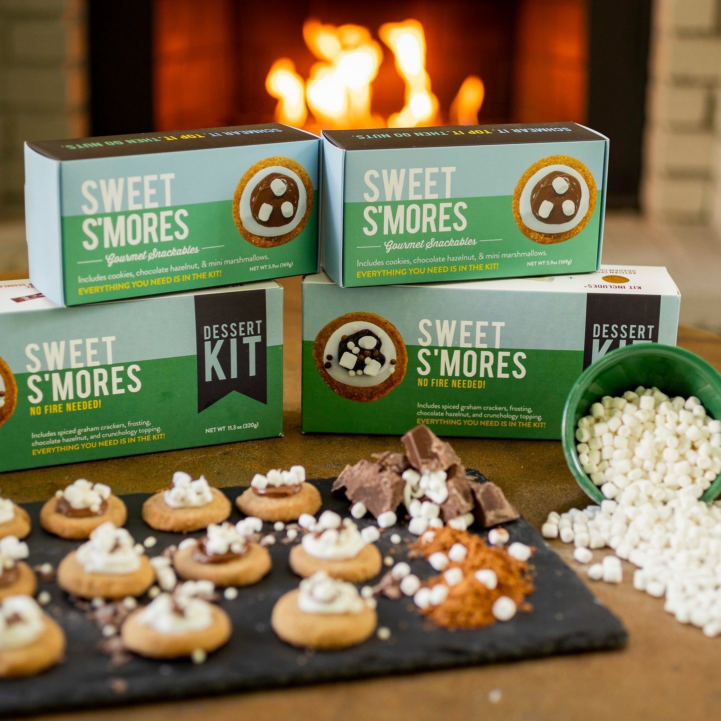 Sweet s'mores dessert kit with marshmallows and chocolate on a table in front of a fireplace.